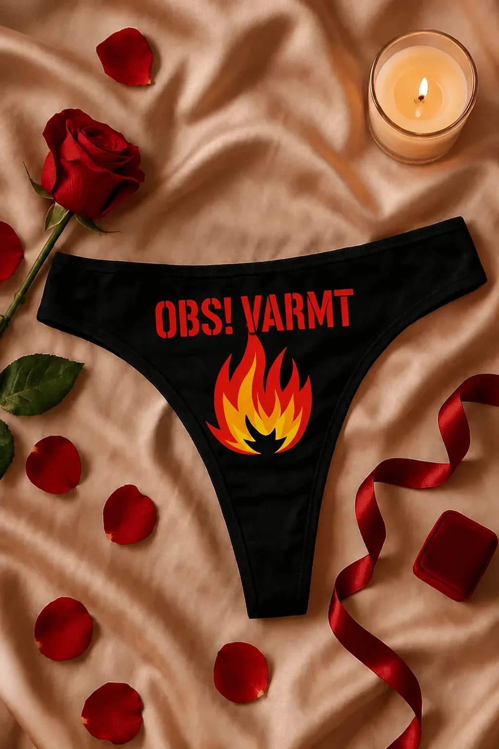 Svart morsom stringtruse med rød tekst og fargerike flammer, liggende blant roser og røde bånd