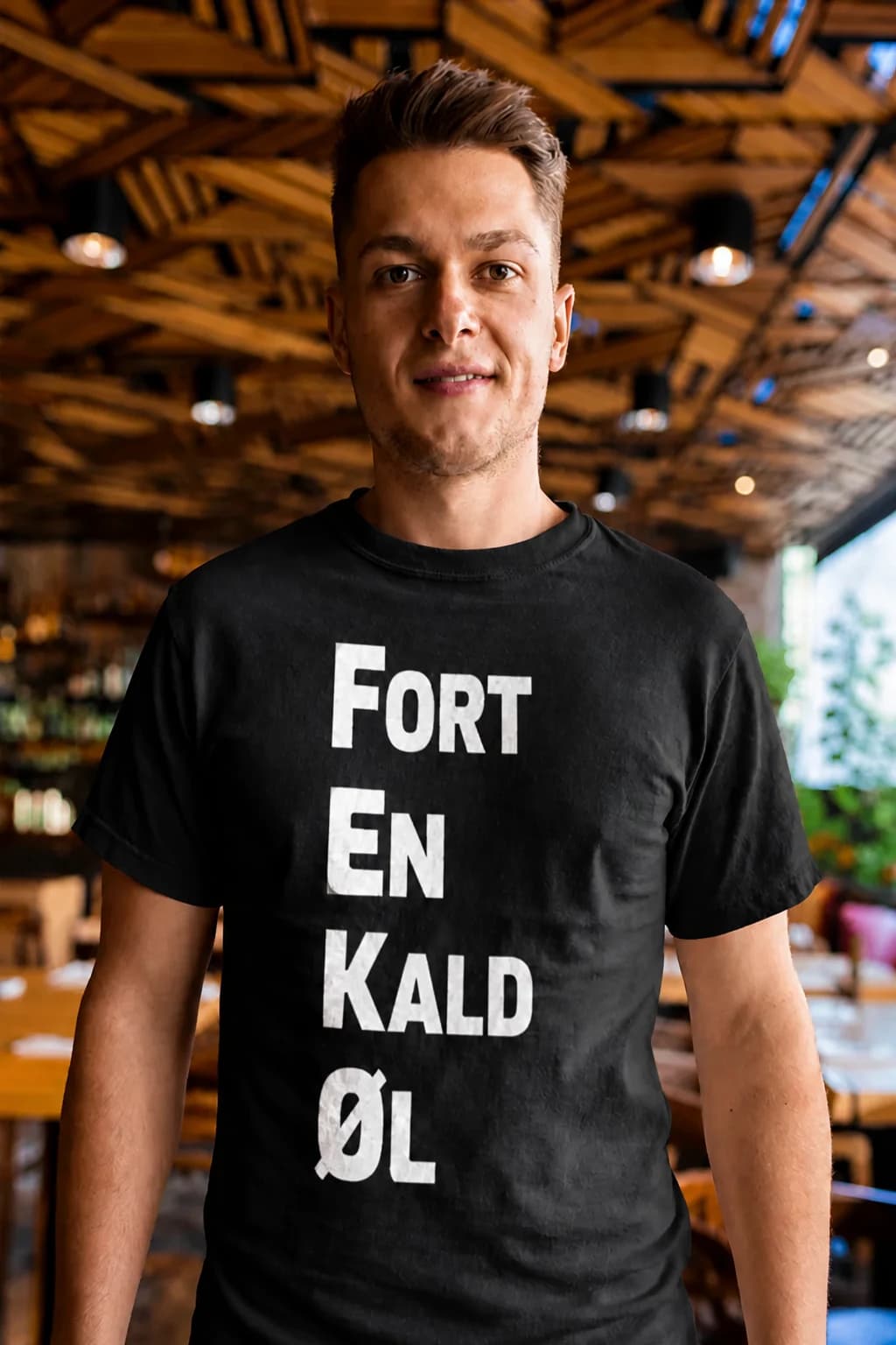 Svart morsom t-skjorte med store hvite bokstaver «Fort en kald øl» på en mann i en pub