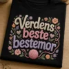 T-skjorte med teksten Verdens beste bestemor i store strikkede bokstaver, med garnnøste og små blomster i varme pastellfarger.
